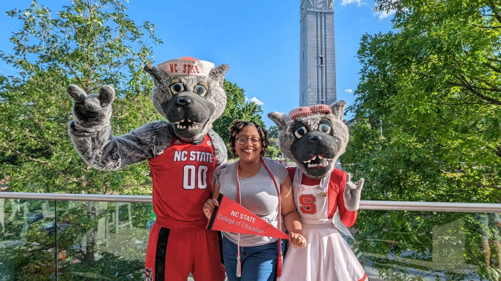 A woman smiling between two NC State mascots outdoors with the NC State belltower in the background.