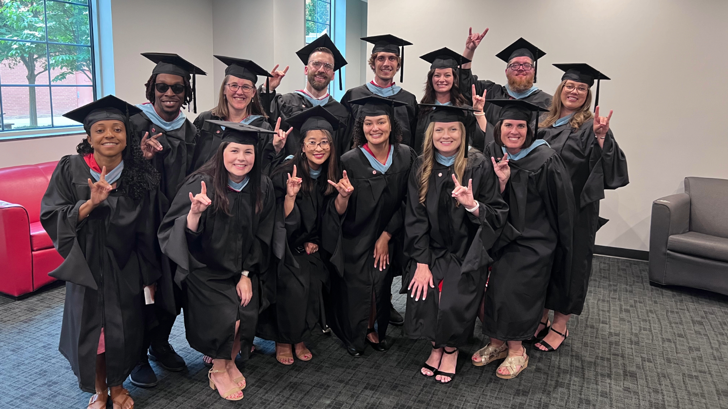 A group of 14 people posing indoors in graduation regalia.