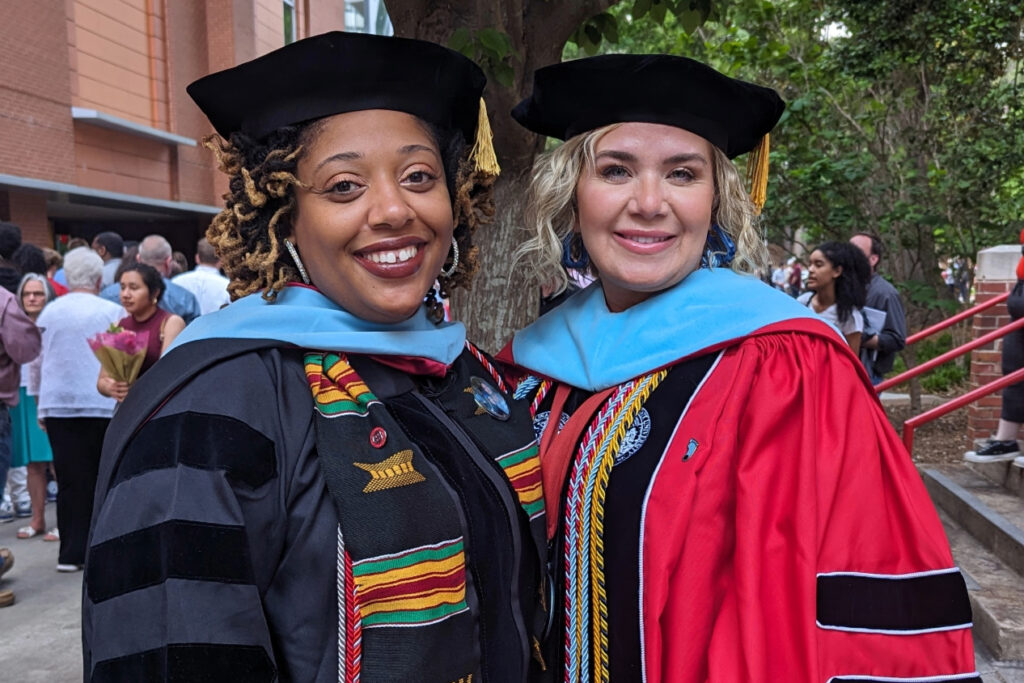 Two women in graduation regalia pose outdoors